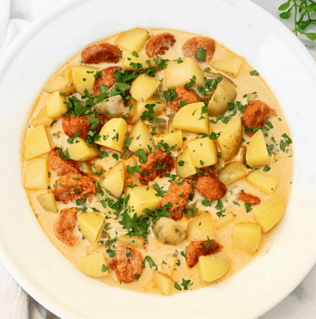 A creamy stew with chunks of potato, pieces of sausage, and sliced mushrooms, garnished with chopped fresh parsley, served in a white bowl.