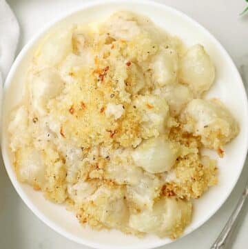 A white bowl filled with creamed onions, topped with a golden, crispy breadcrumb crust, sits on a light surface.