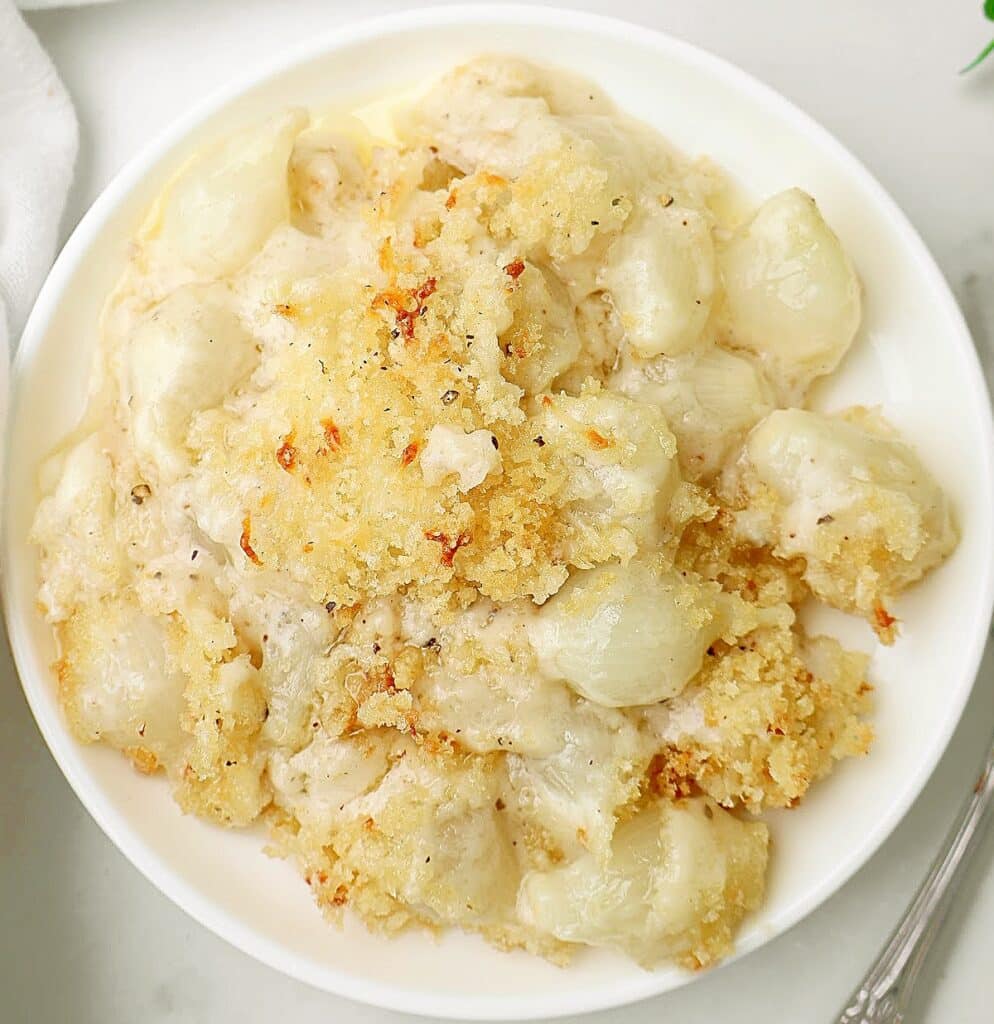 A white bowl filled with creamed onions, topped with a golden, crispy breadcrumb crust, sits on a light surface.