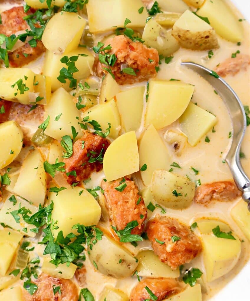 A close-up of a creamy stew with diced potatoes, sausage pieces, chopped parsley, and a spoon partially visible on the right side.