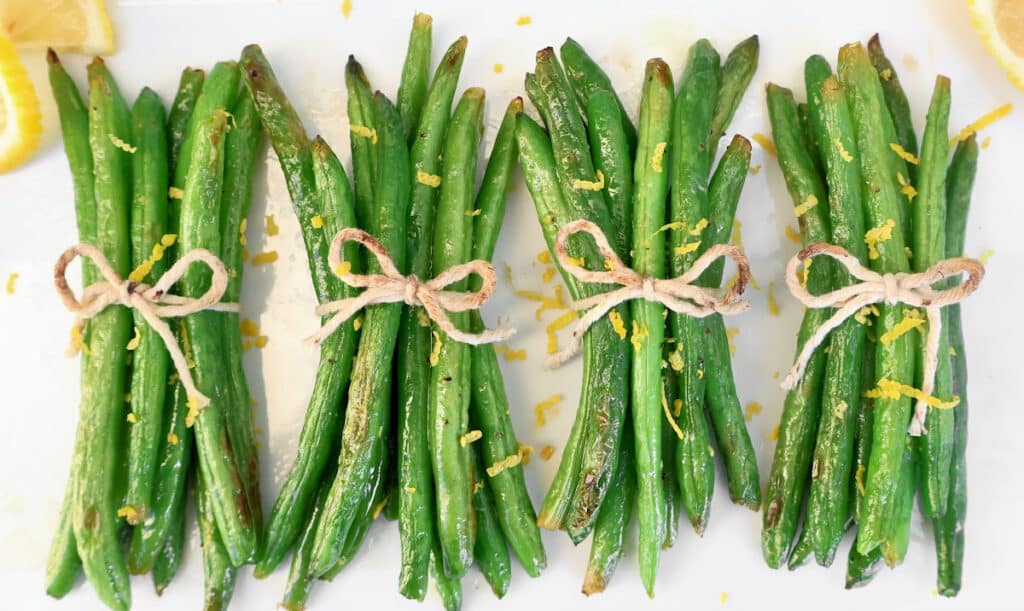 Four green bean bundles tied with twine are arranged in a row on a white surface, topped with lemon zest and flanked by fresh lemon wedges.