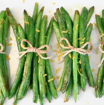 Four green bean bundles tied with twine are arranged in a row on a white surface, topped with lemon zest and flanked by fresh lemon wedges.