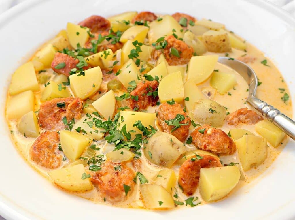A creamy soup with chunks of potatoes, sliced sausage, and garnished with chopped parsley, served in a white bowl with a spoon.