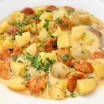 A creamy soup with chunks of potatoes, sliced sausage, and garnished with chopped parsley, served in a white bowl with a spoon.