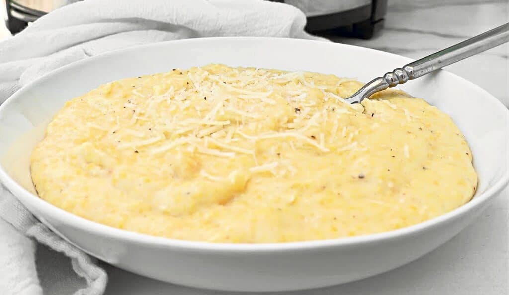A white bowl filled with creamy yellow grits, topped with shredded cheese, with a silver spoon resting inside. A white cloth is placed beside the bowl on a light-colored surface.