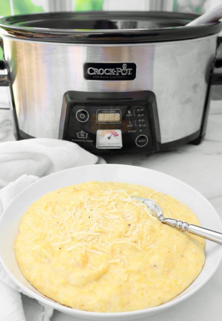 A bowl of creamy polenta topped with shredded cheese sits in front of a stainless steel Crock-Pot slow cooker on a white counter. A fork rests in the bowl.