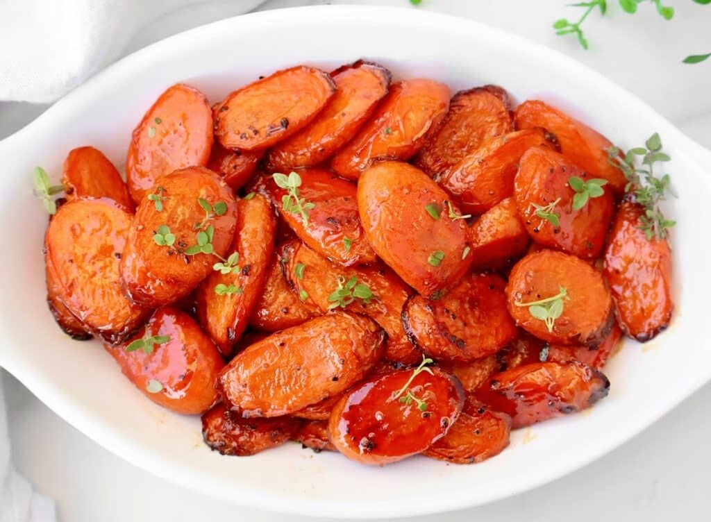 A white oval dish filled with balsamic roasted carrots, glazed and golden brown, garnished with fresh green herbs. The roasted carrot slices are arranged neatly in the dish.