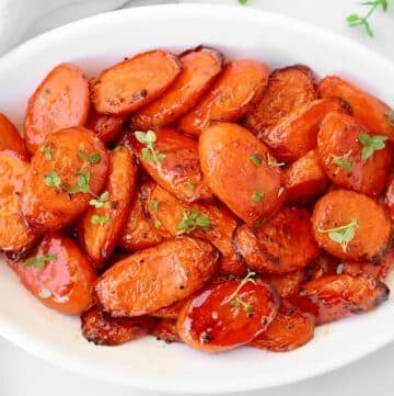 A white oval dish filled with balsamic roasted carrots, glazed and golden brown, garnished with fresh green herbs. The roasted carrot slices are arranged neatly in the dish.