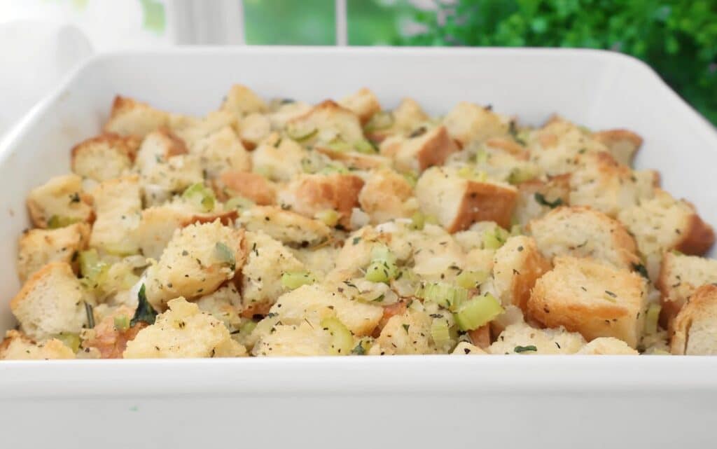 A white baking dish filled with homemade bread stuffing, featuring chunks of bread, diced celery, herbs, and seasoning, with a blurred green background.