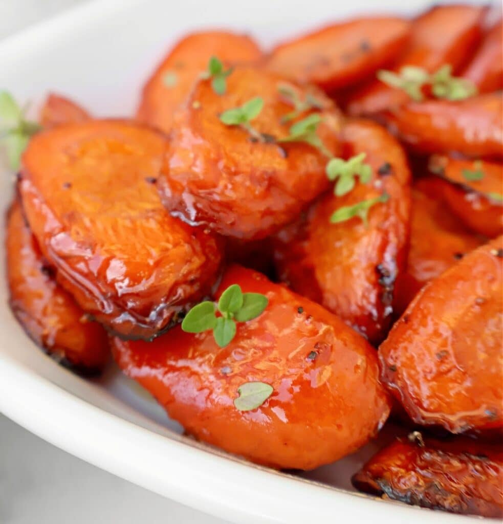Close-up of balsamic roasted carrots glazed with a shiny coating, garnished with small sprigs of fresh green herbs, served in a white dish.