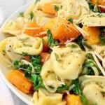 A close-up of tortellini with butternut squash, sautéed kale, grated parmesan cheese, and fresh herbs. The colorful dish is garnished with cracked black pepper for a vibrant finish.