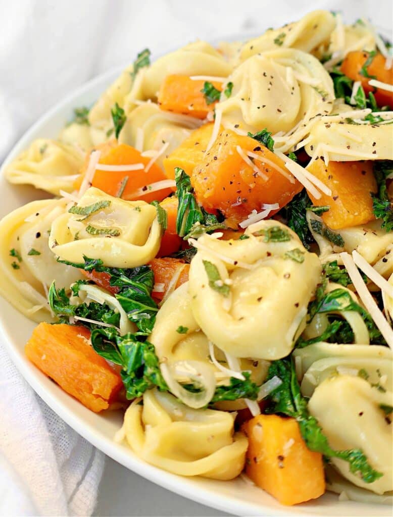 A close-up of tortellini with butternut squash, sautéed kale, grated parmesan cheese, and fresh herbs. The colorful dish is garnished with cracked black pepper for a vibrant finish.