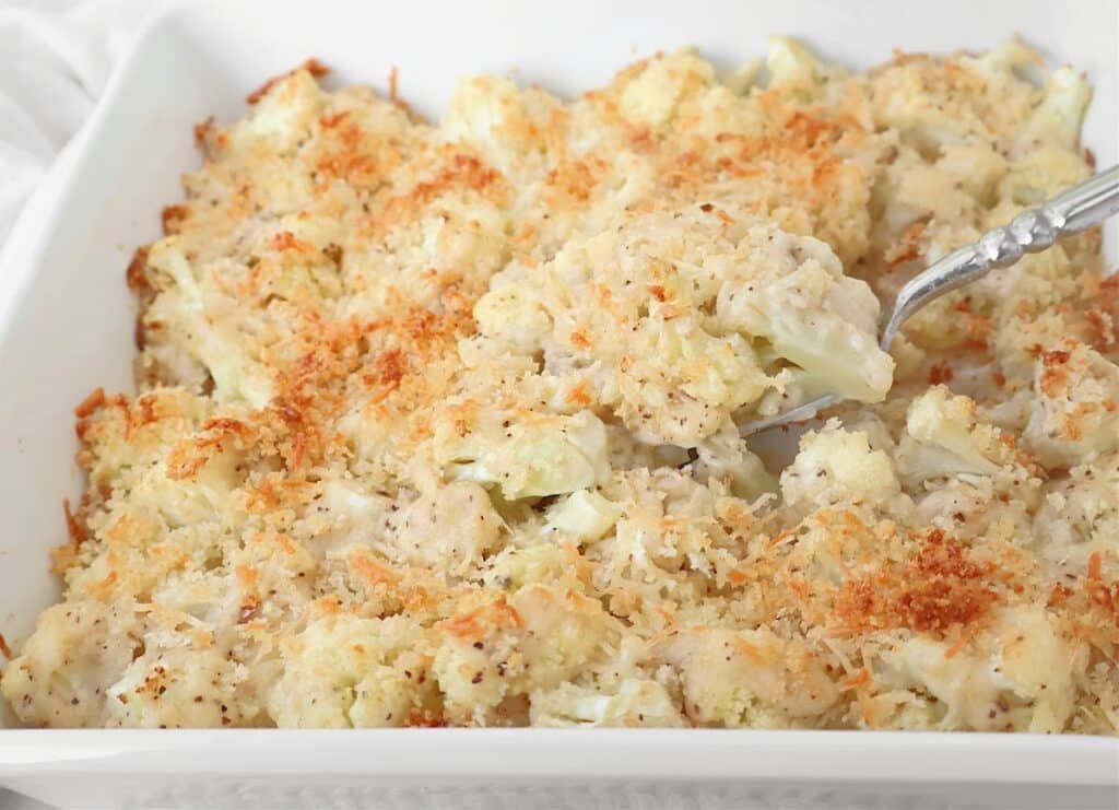 A close-up of a roasted cauliflower gratin in a white dish, topped with golden brown breadcrumbs. A serving spoon is scooping a portion of the creamy, cheesy cauliflower mixture.