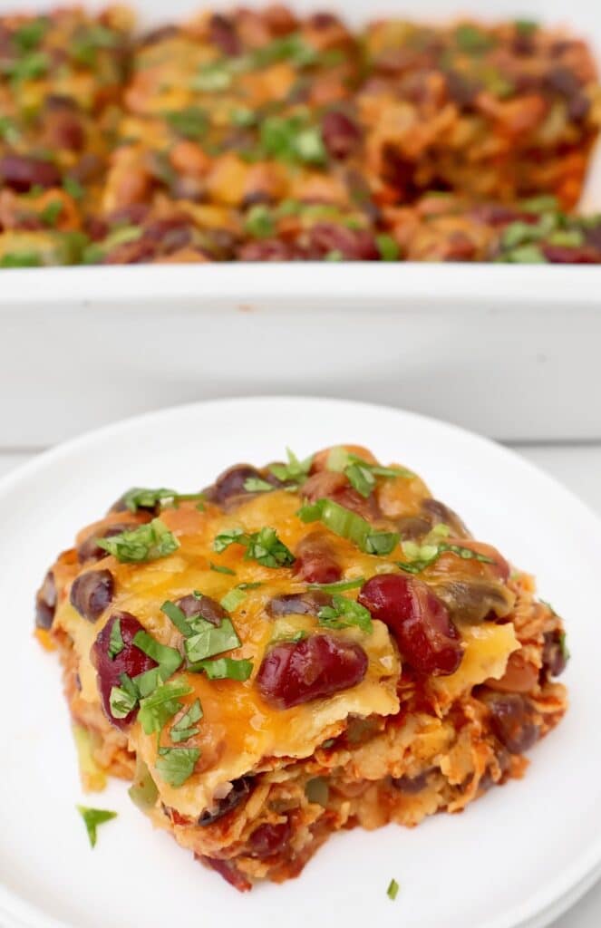 A serving of cheesy bean enchilada casserole filled with beans, vegetables, and topped with melted cheese and chopped cilantro sits on a white plate, with the remaining casserole in a dish in the background.