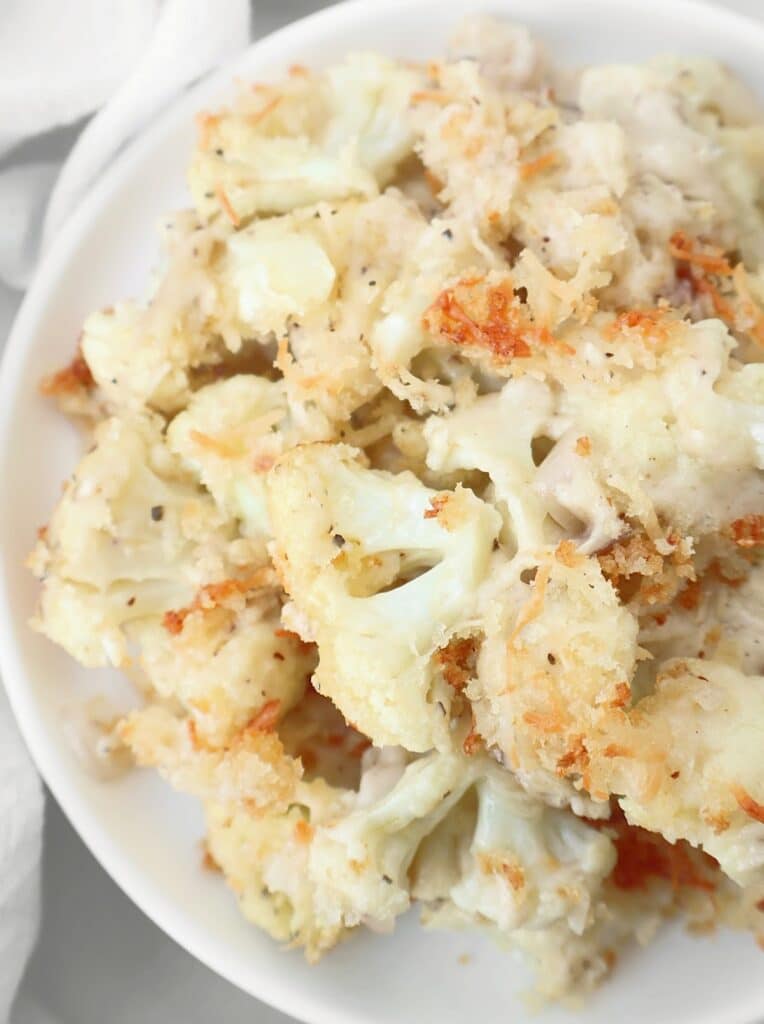 A close-up of roasted cauliflower gratin features florets coated in a creamy sauce, topped with golden, crispy breadcrumbs and melted cheese, served on a white plate.