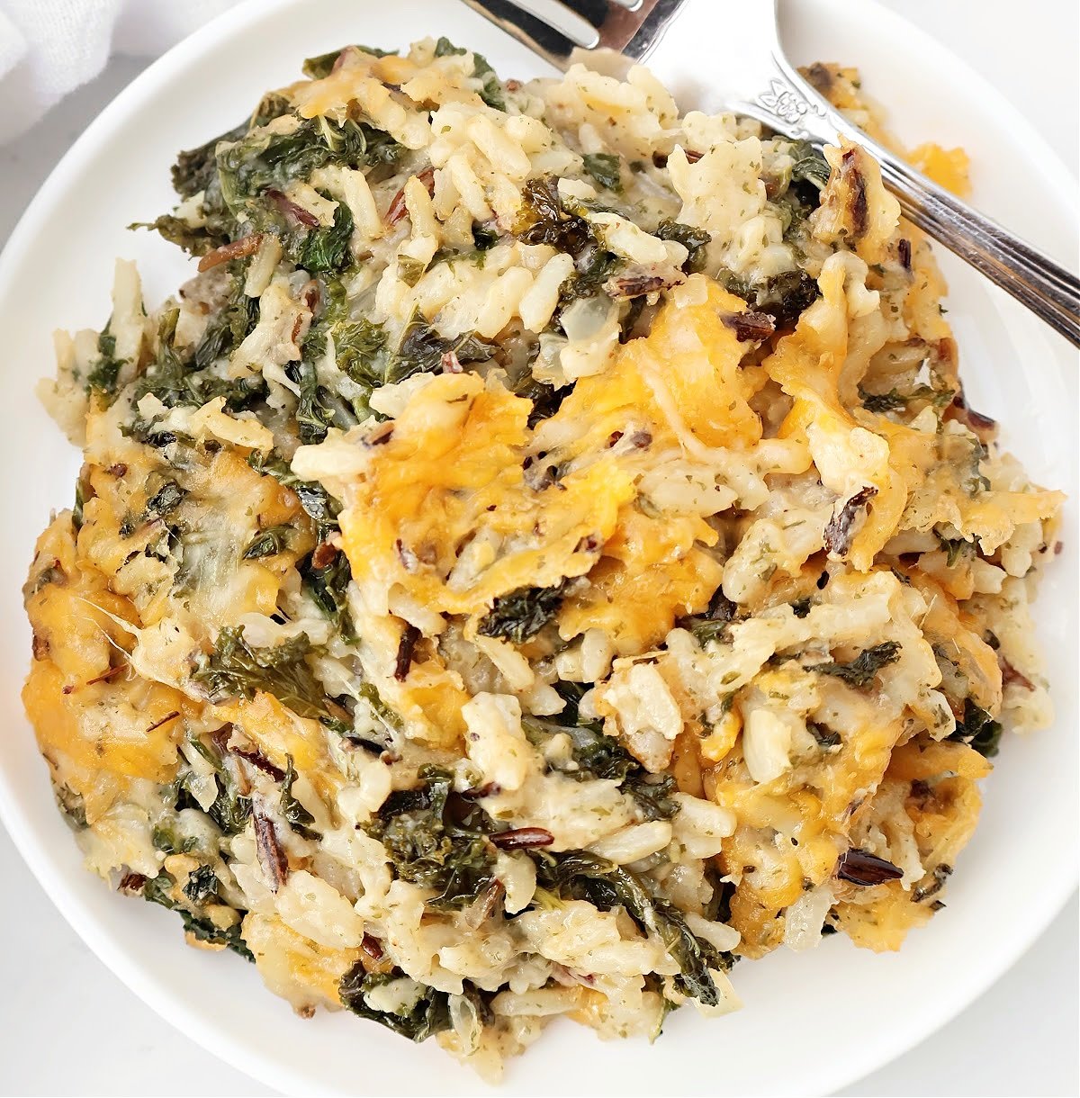 A close-up of a plate with a cheesy, baked casserole featuring wild rice, leafy greens, and melted cheese, with a fork on the side.