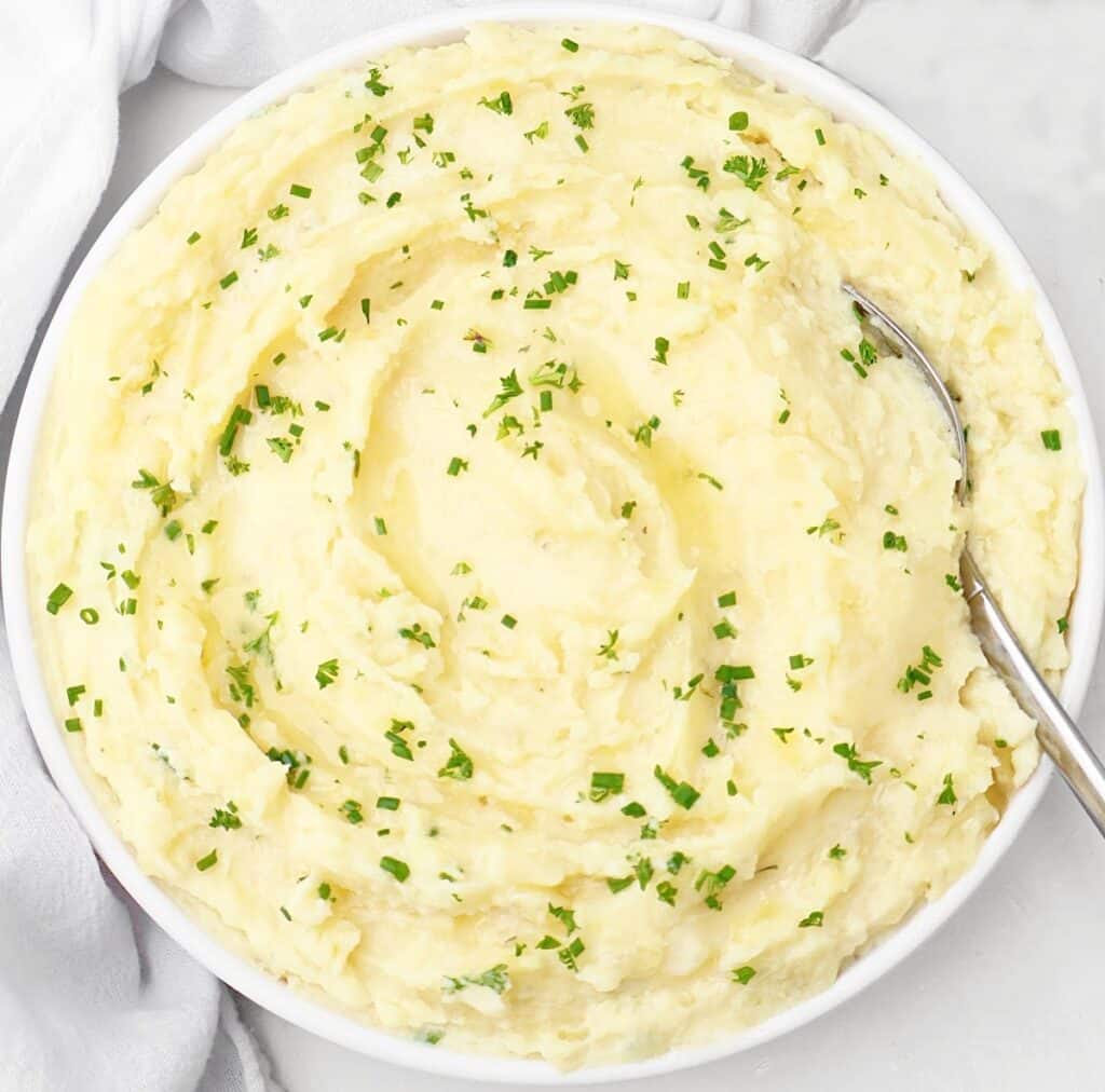 A bowl of creamy mashed potatoes topped with chopped fresh herbs, with a spoon resting on the side of the bowl.
