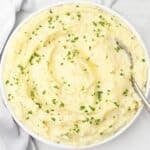 A bowl of creamy mashed potatoes topped with chopped fresh herbs, with a spoon resting on the side of the bowl.