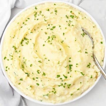 A bowl of creamy mashed potatoes topped with chopped fresh herbs, with a spoon resting on the side of the bowl.