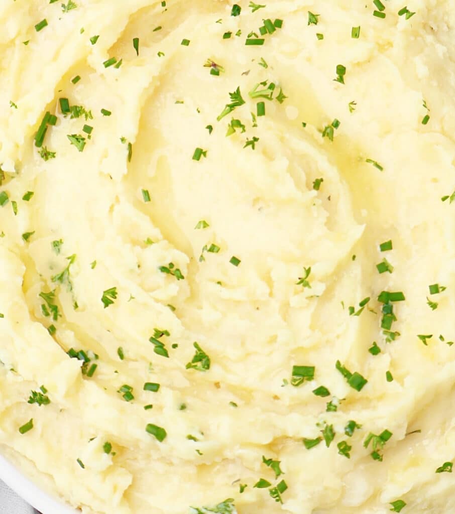 Creamy mashed potatoes garnished with finely chopped green herbs, served in a white dish. The texture appears smooth and fluffy with a few small lumps.
