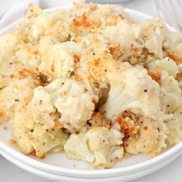 A white plate filled with roasted cauliflower gratin, featuring baked florets topped with a golden, crispy breadcrumb and cheese coating. A fork is partially visible in the background.