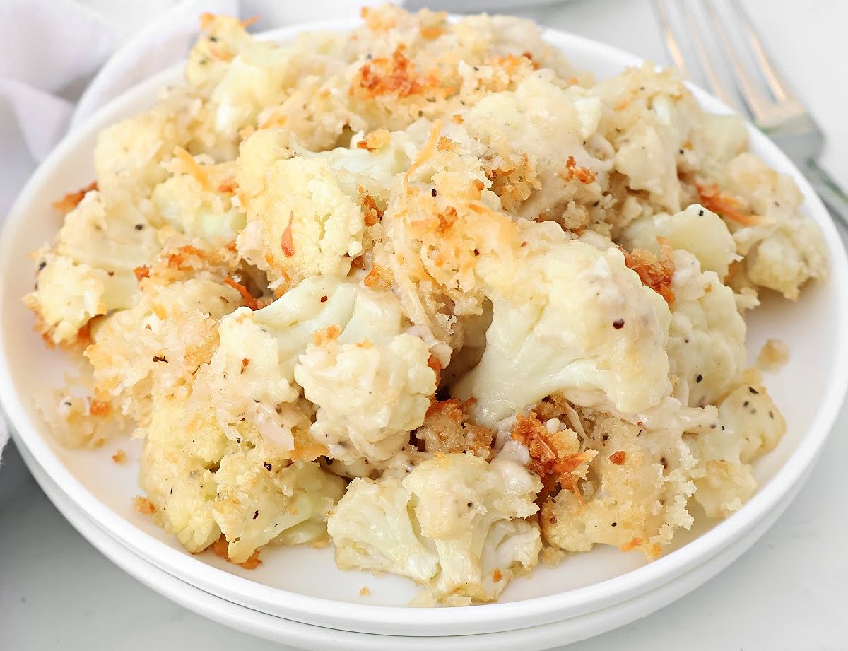 A white plate filled with roasted cauliflower gratin, featuring baked florets topped with a golden, crispy breadcrumb and cheese coating. A fork is partially visible in the background.