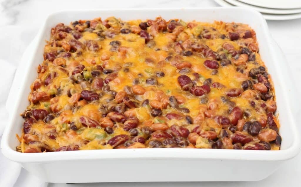 A white square casserole dish filled with a bean enchilada casserole, baked with chili, melted cheddar cheese, kidney beans, pinto beans, and green peppers. Plates are stacked in the background.