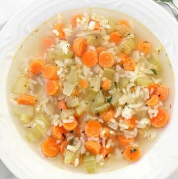 A bowl of clear vegetable soup with sliced carrots, celery, and rice, served in a white bowl on a white surface. A silver spoon is partially visible in the top right corner.