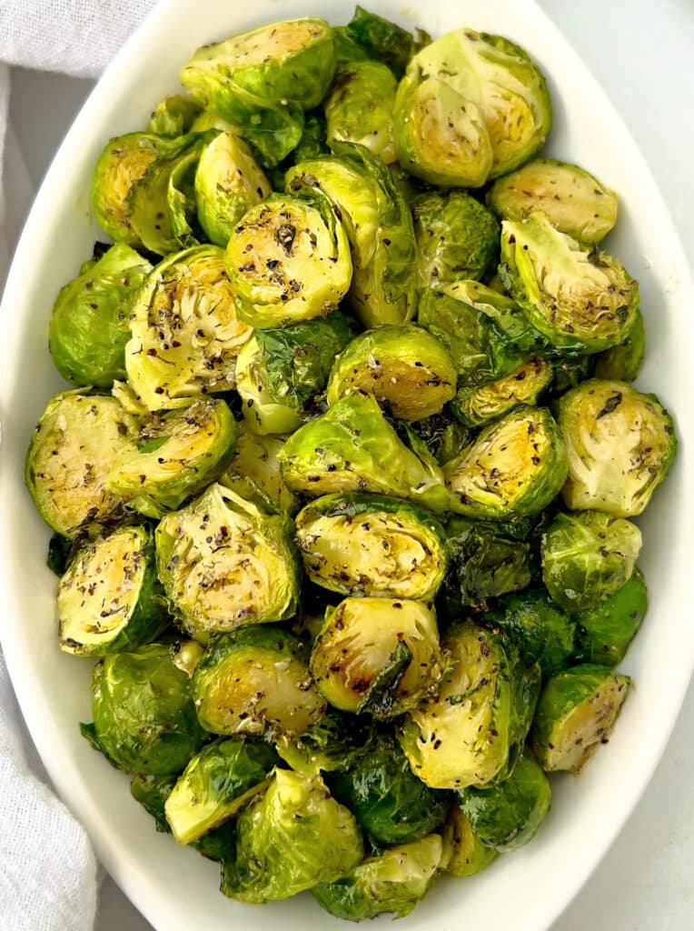 A white oval dish filled with roasted Brussels sprouts, halved and seasoned with herbs and spices, garnished with flecks of black pepper.