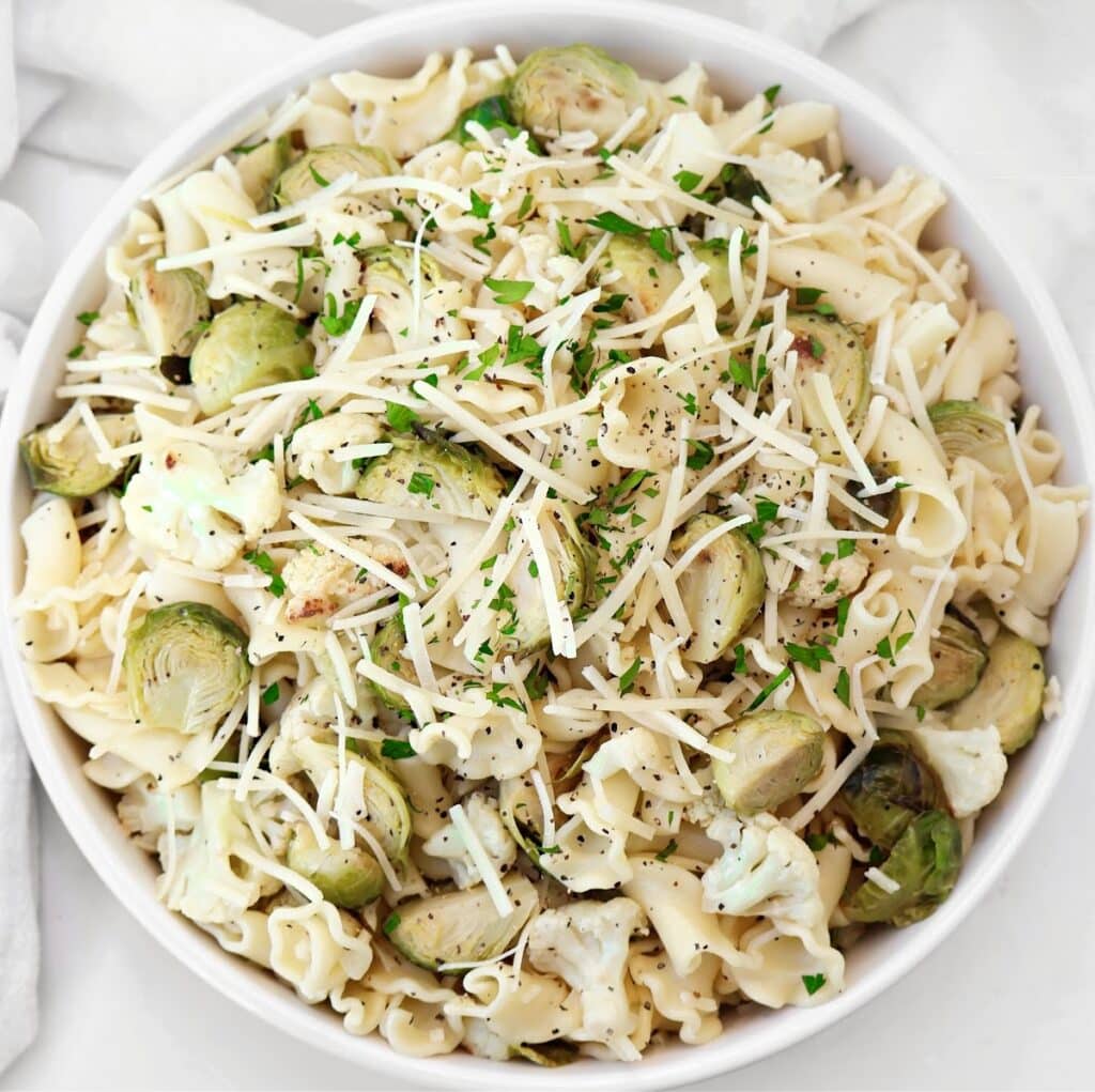 A bowl of roasted vegetable pasta salad topped with Brussels sprouts, cauliflower, shredded cheese, chopped parsley, and black pepper.