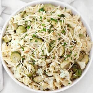 A bowl of roasted vegetable pasta salad topped with Brussels sprouts, cauliflower, shredded cheese, chopped parsley, and black pepper.