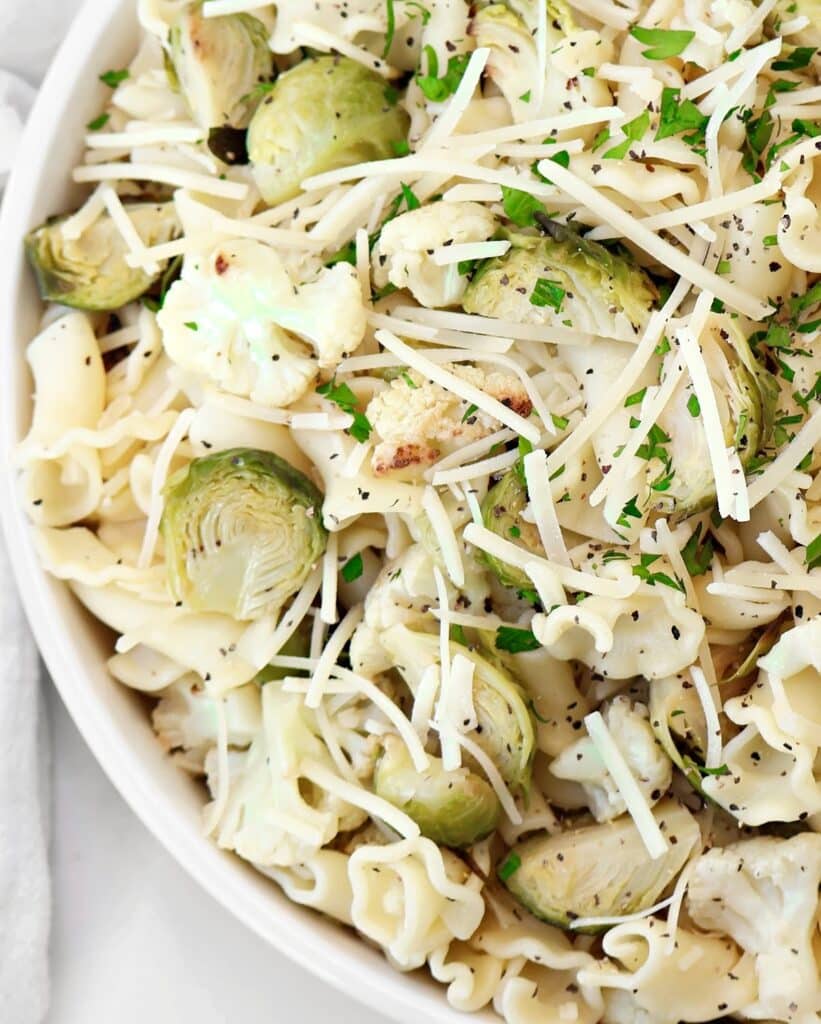 Roasted vegetable pasta salad featuring pasta topped with roasted Brussels sprouts, cauliflower, shredded Parmesan cheese, and chopped fresh herbs, served in a white bowl.