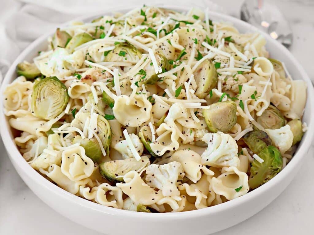 A white bowl filled with roasted vegetable pasta salad featuring pasta, Brussels sprouts, cauliflower florets, shredded cheese, and chopped herbs, with a spoon partially visible in the background.