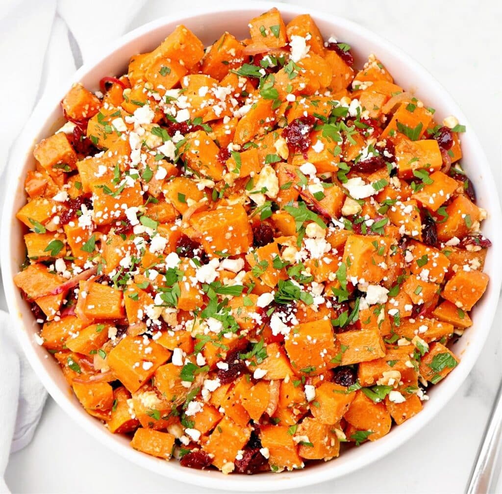 A white bowl filled with roasted sweet potato salad, garnished with crumbled cheese, chopped herbs, and small bits of dried cranberries on a white surface.