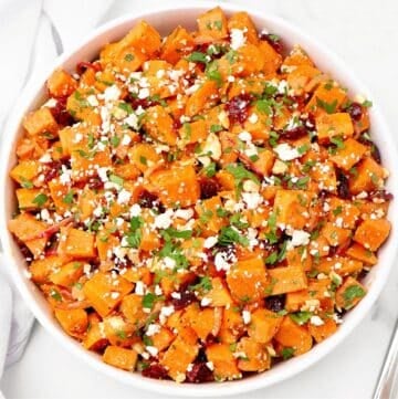 A white bowl filled with roasted sweet potato salad, garnished with crumbled cheese, chopped herbs, and small bits of dried cranberries on a white surface.