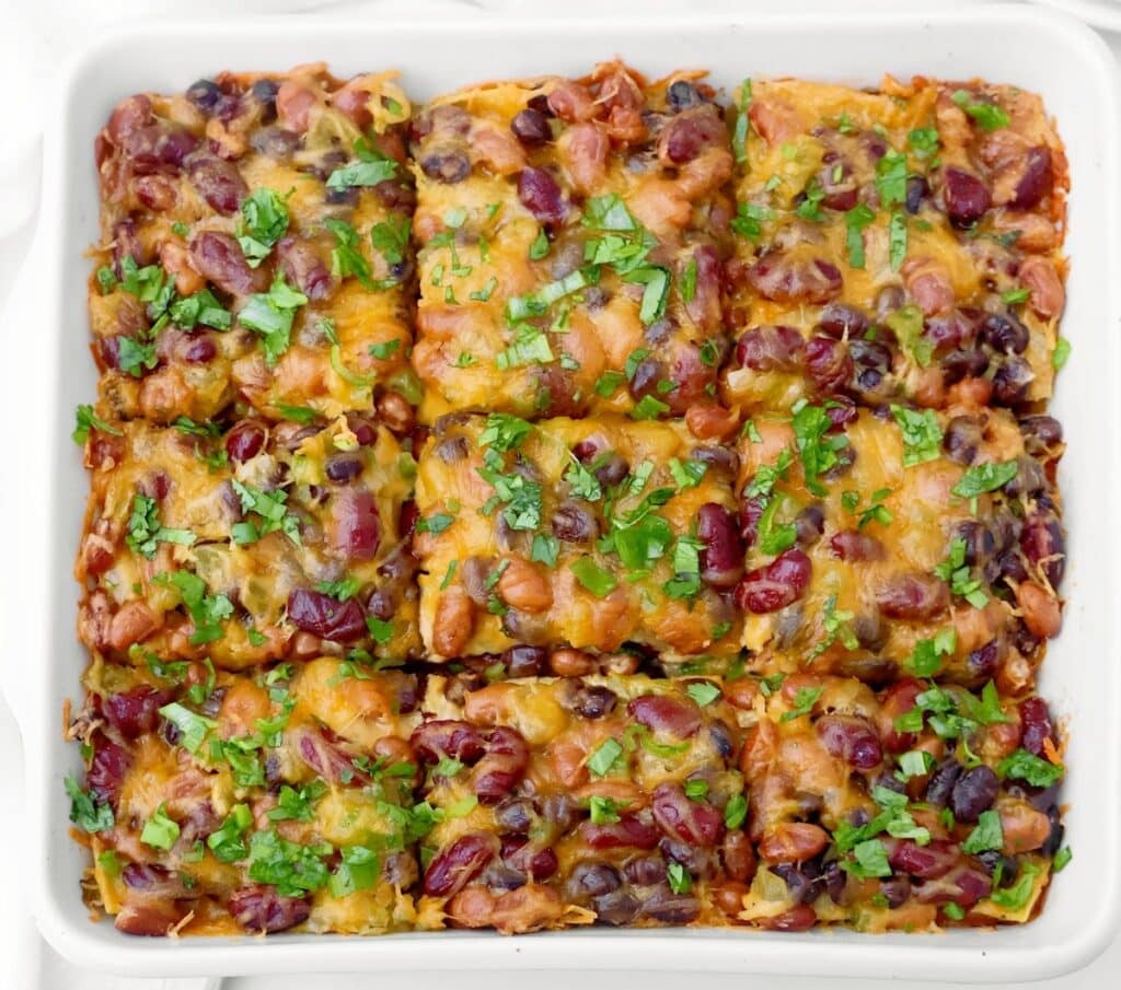 Baked bean enchilada casserole cut into nine squares, topped with melted cheese, red kidney beans, black beans, and fresh herbs, served in a white square dish.