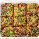 Baked bean enchilada casserole cut into nine squares, topped with melted cheese, red kidney beans, black beans, and fresh herbs, served in a white square dish.