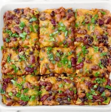 Baked bean enchilada casserole cut into nine squares, topped with melted cheese, red kidney beans, black beans, and fresh herbs, served in a white square dish.