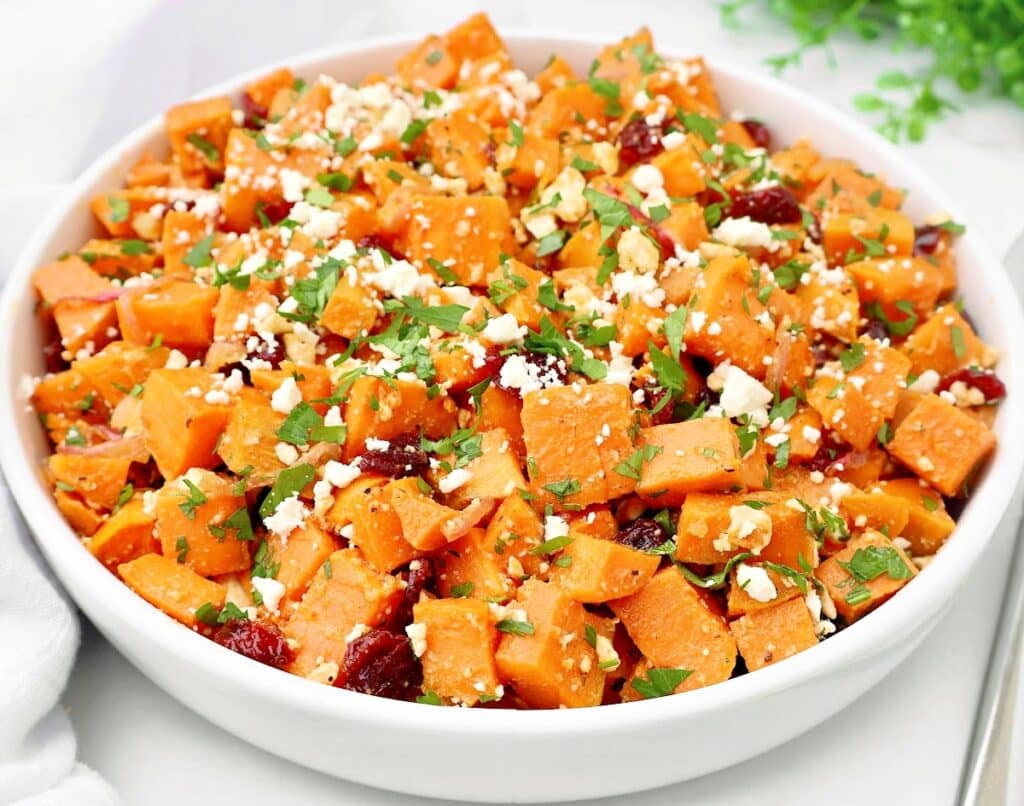 A vibrant roasted sweet potato salad in a white bowl, garnished with chopped parsley, crumbled feta cheese, and dried cranberries. The colorful dish sits beautifully on a white surface.