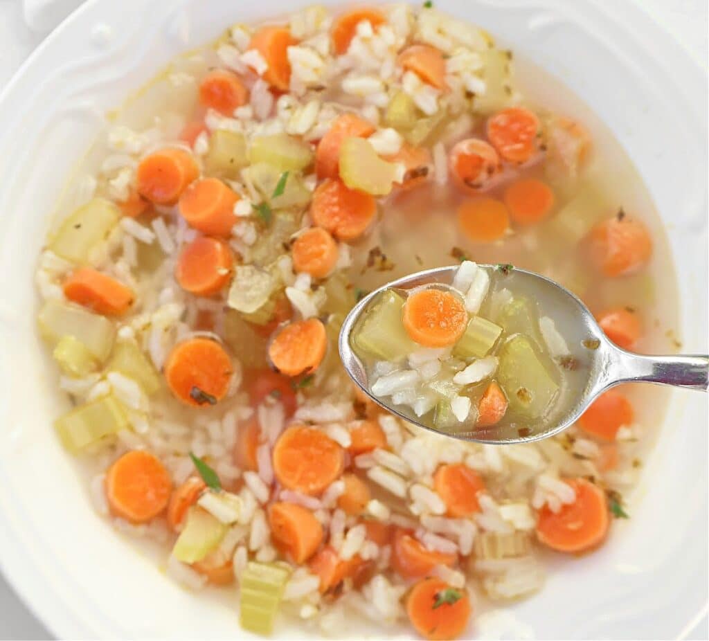 A bowl of soup with sliced carrots, celery, rice, and herbs, with a spoonful of the soup held above the bowl.