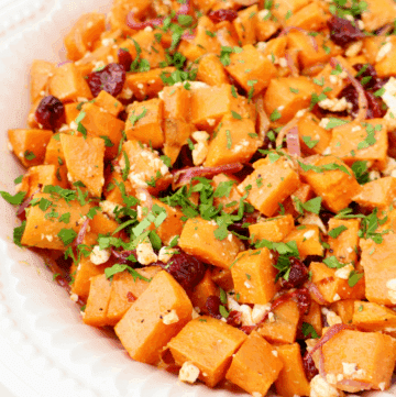 A close-up of a roasted sweet potato salad featuring roasted sweet potato cubes, dried cranberries, crumbled feta cheese, sliced red onions, and chopped fresh parsley served in a white scalloped dish.