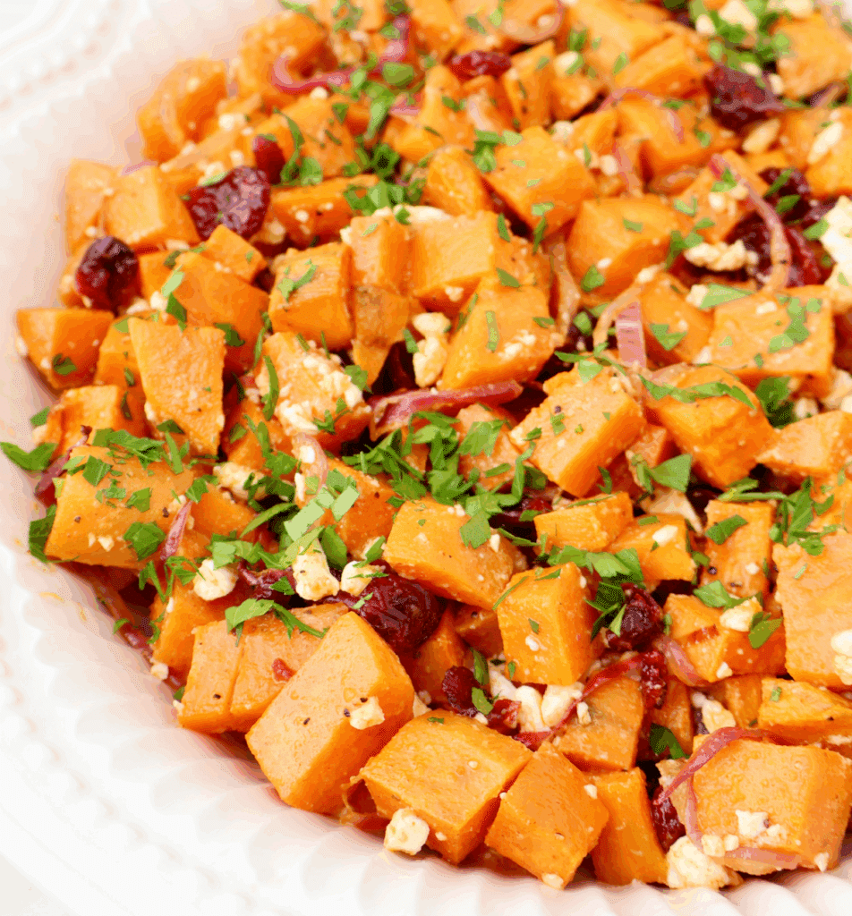 A close-up of a roasted sweet potato salad featuring roasted sweet potato cubes, dried cranberries, crumbled feta cheese, sliced red onions, and chopped fresh parsley served in a white scalloped dish.