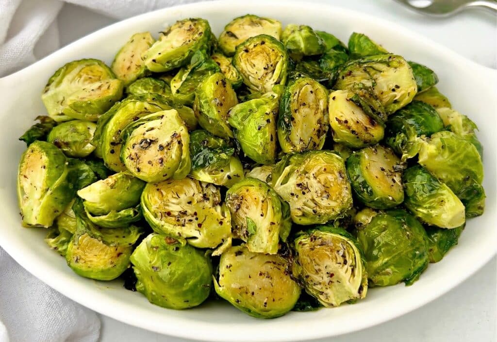 A white oval dish filled with roasted Brussels sprouts, cut in halves and seasoned with herbs and spices, sits on a white surface.