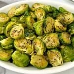 A white oval dish filled with roasted Brussels sprouts, cut in halves and seasoned with herbs and spices, sits on a white surface.