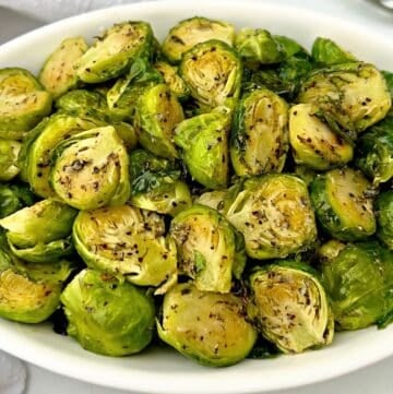 A white oval dish filled with roasted Brussels sprouts, cut in halves and seasoned with herbs and spices, sits on a white surface.