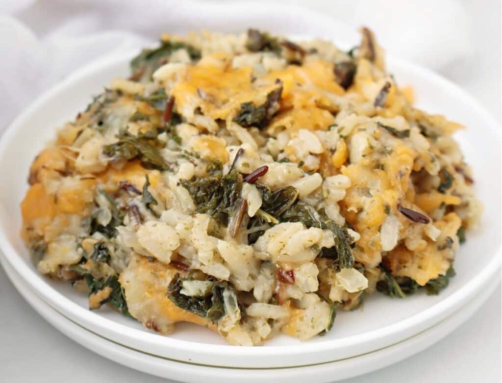 A close-up of a serving of cheesy wild rice casserole with spinach, cheese, and a creamy sauce on a white plate.