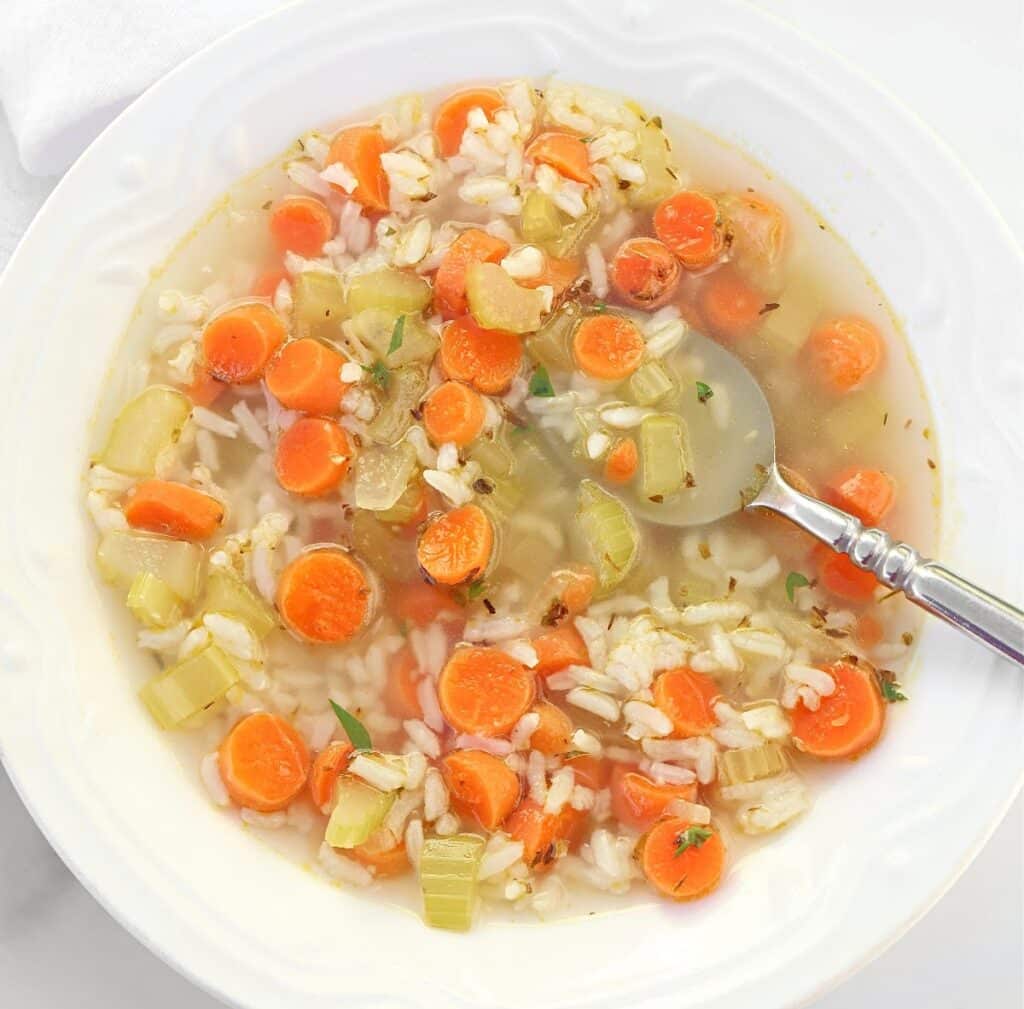 A bowl of vegetable soup with sliced carrots, celery, rice, and herbs in a clear broth. A spoon rests in the bowl.