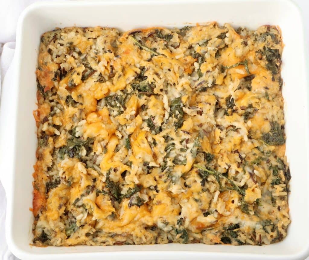 A white baking dish filled with a baked casserole made of rice, cheese, and leafy greens. The casserole has a golden-brown top with visible bits of melted cheese and green herbs mixed throughout.