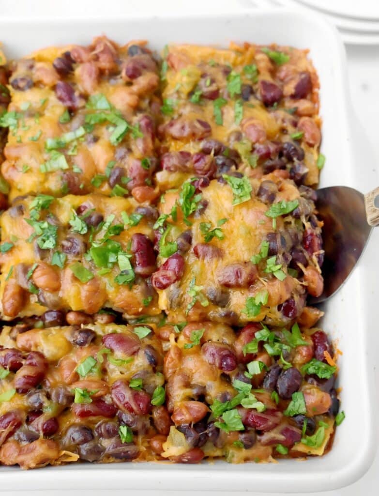 A bean enchilada casserole filled with baked enchiladas, topped with melted cheese, kidney beans, black beans, and fresh cilantro. A serving spatula lifts one portion from the dish.