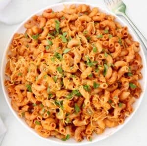 A white bowl filled with American goulash—macaroni pasta in a rich red tomato sauce, topped with melted cheese and chopped fresh parsley—sits on a white surface with a fork placed nearby.